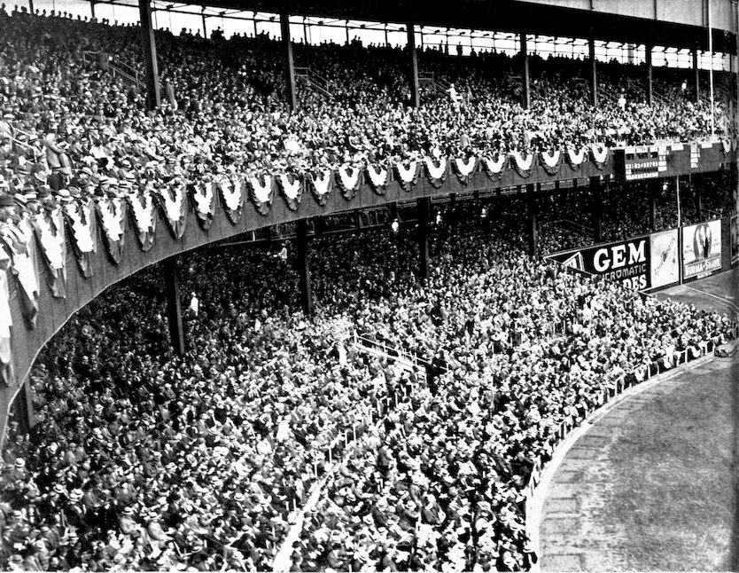 A massive, crowded baseball stadium grandstand from the early-to-mid 20th century, filled with spectators wearing hats.