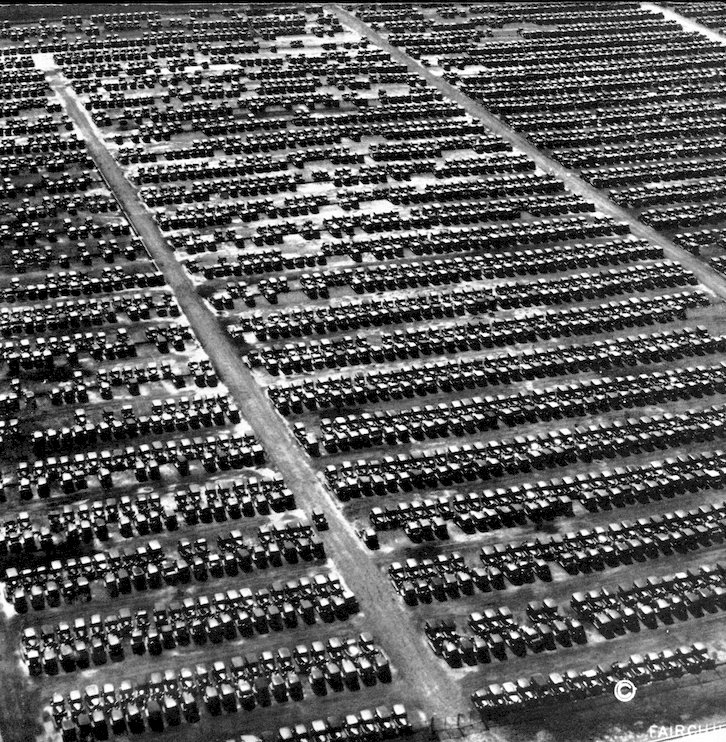 An aerial, black and white photograph showing thousands of vintage automobiles parked in tight, orderly rows across a massive outdoor lot.