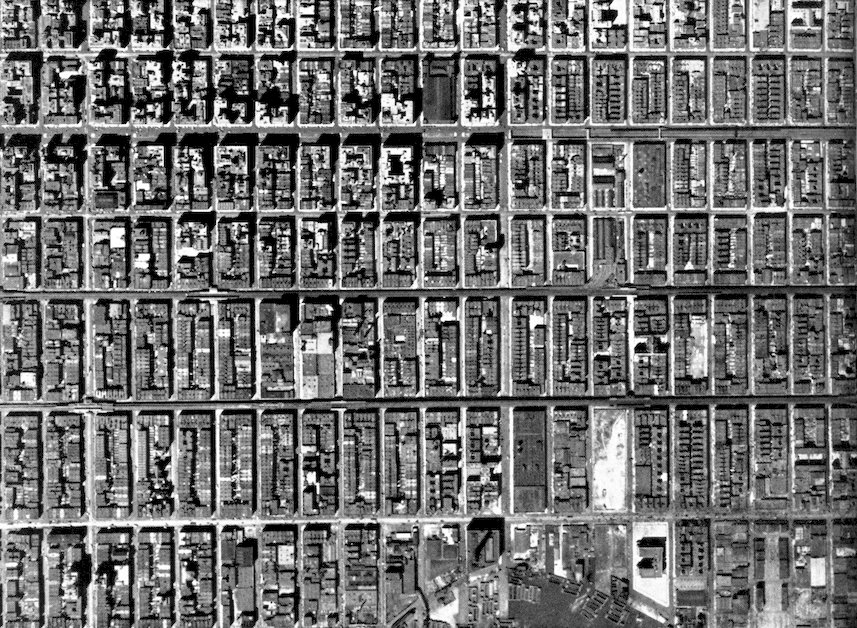 A high-angle, black and white aerial photograph showing a dense, rigid grid of urban city blocks and buildings with long afternoon shadows.