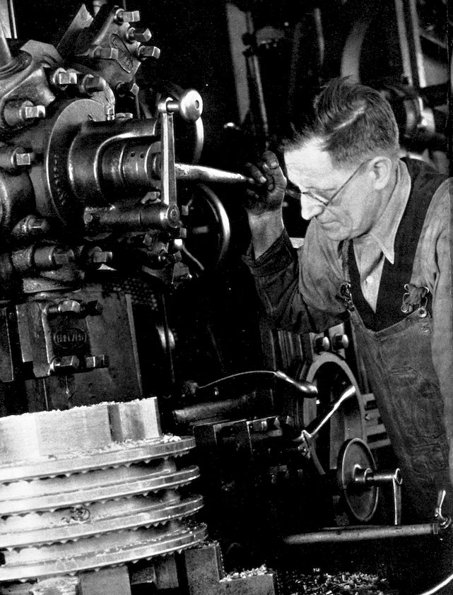 A black and white historical photograph of a machinist wearing glasses and overalls, carefully operating a large industrial lathe or turret machinery.
