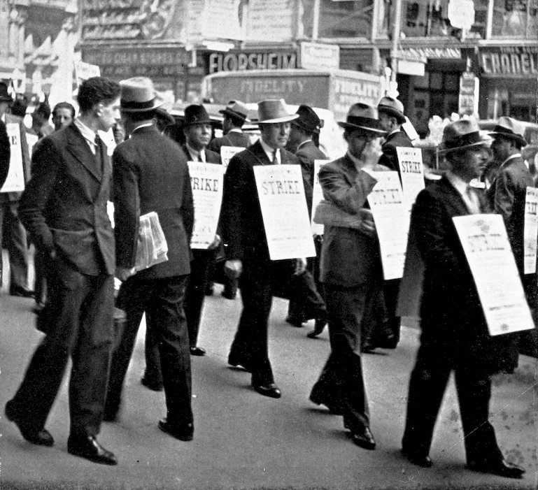 A historical black and white photograph of a labor strike, showing men in suits and fedoras walking along a city street wearing sandwich-board signs that read 'STRIKE'.